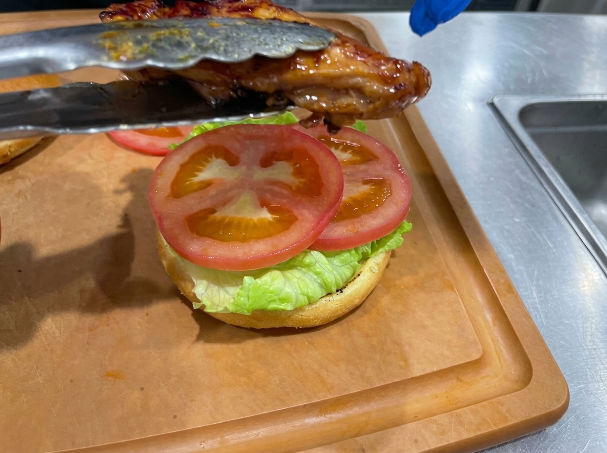 Una sovracoscia di pollo teriyaki appena cotta posizionata con le pinze sopra un panino farcito con lattuga e pomodoro.