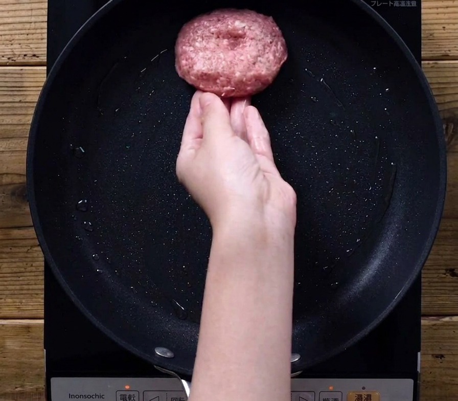 Una mano che adagia la polpetta di carne in una padella nera su un fornello a induzione.