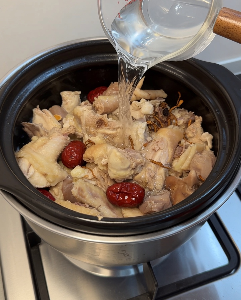 Acqua limpida versata da un misurino in una pentola di coccio con pollo, datteri ed erbe.