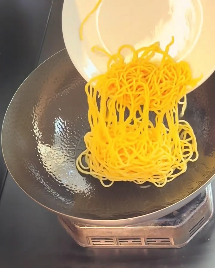 Spaghetti gialli cotti che scivolano da un piatto bianco in un wok caldo oliato.