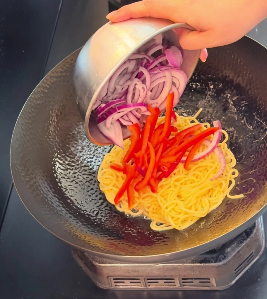 Vista ravvicinata di straccetti di manzo, strisce di peperone rosso e fette di cipolla versate nel wok con gli spaghetti.