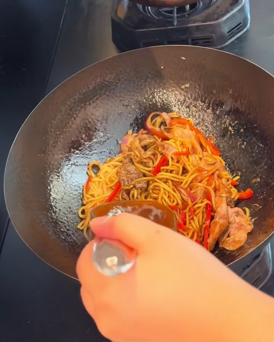 Una mano che usa un cucchiaio per saltare gli spaghetti al pepe nero in un wok a fiamma alta.