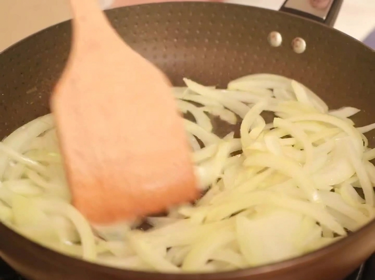 Strisce di cipolla dorata che cuociono in padella con olio, mescolate con una spatola di legno.
