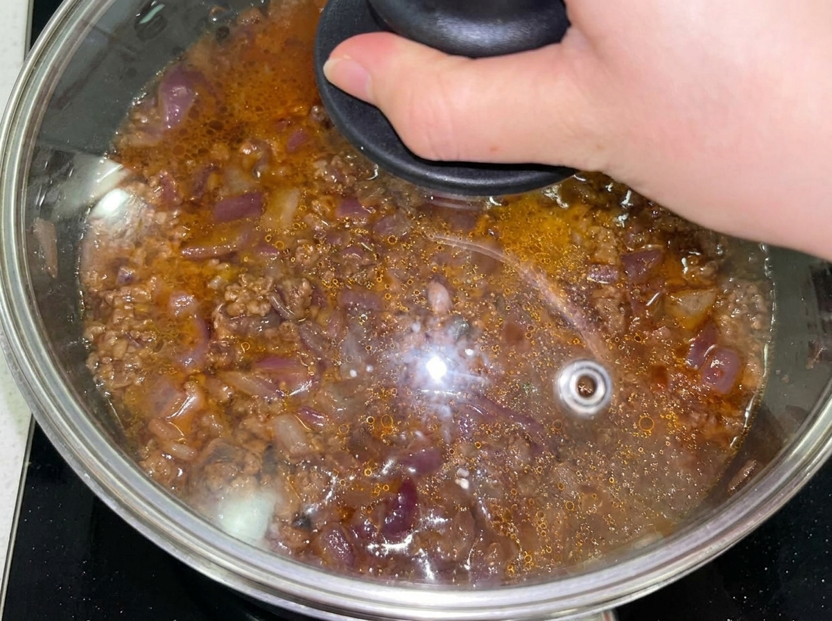 Composto di manzo e cipolla in padella coperto con un coperchio di vetro dopo aver aggiunto acqua.