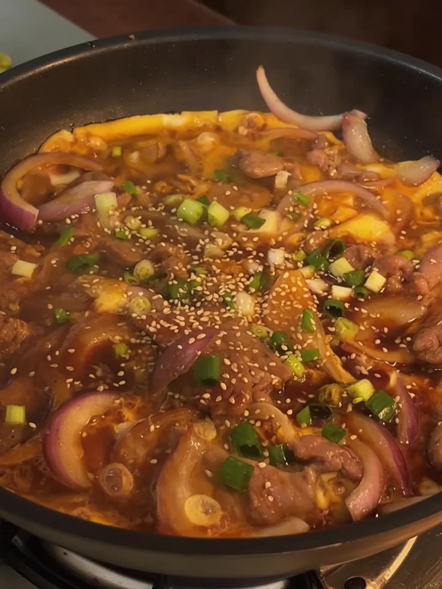 Un piatto finito di manzo e uova in una padella, guarnito con cipollotti freschi tritati e semi di sesamo tostati.