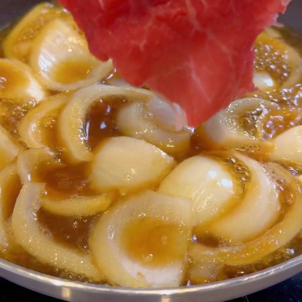 Fettine sottili di manzo crudo disposte in padella con cipolle e salsa scura in ebollizione.