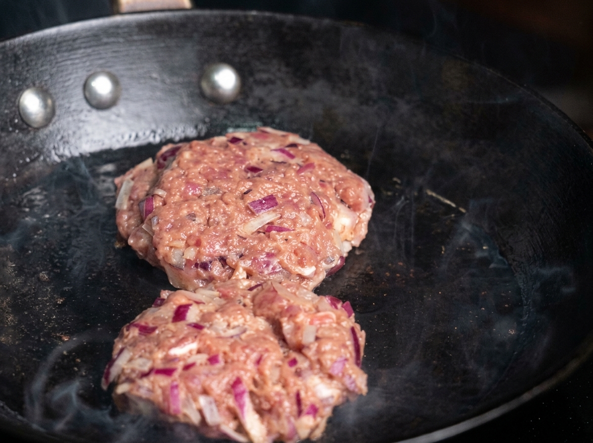Due hamburger crudi conditi posti in una padella calda unta, con fumo visibile.