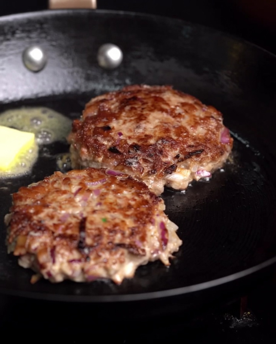 Hamburger in cottura in padella con una noce di burro sciolto e un rametto di rosmarino fresco.