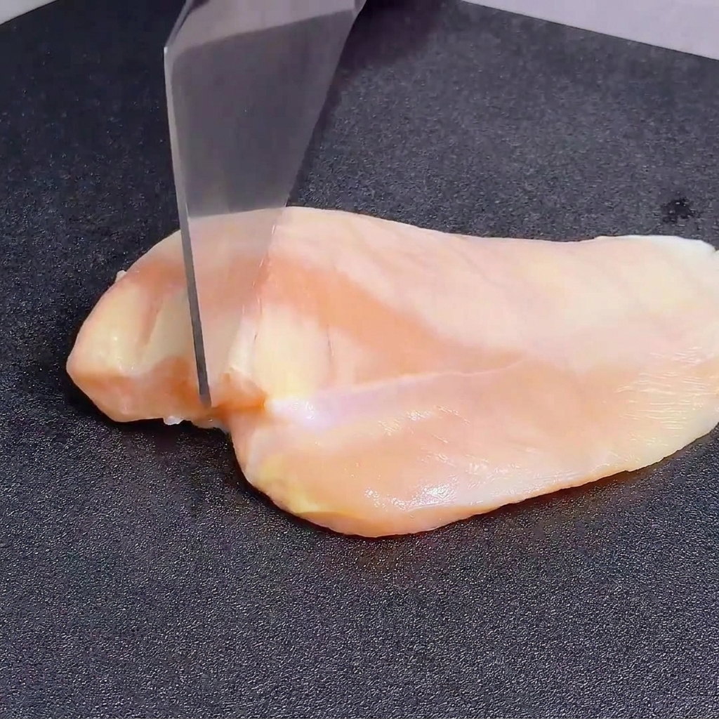 Battere un petto di pollo crudo con il dorso di un grosso coltello su un tagliere scuro.