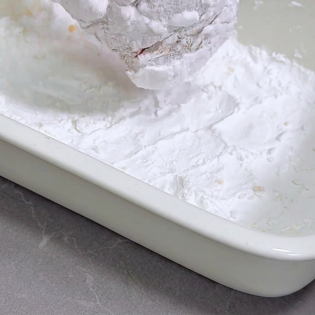 Petto di pollo marinato premuto in una vaschetta bianca piena di amido di mais.