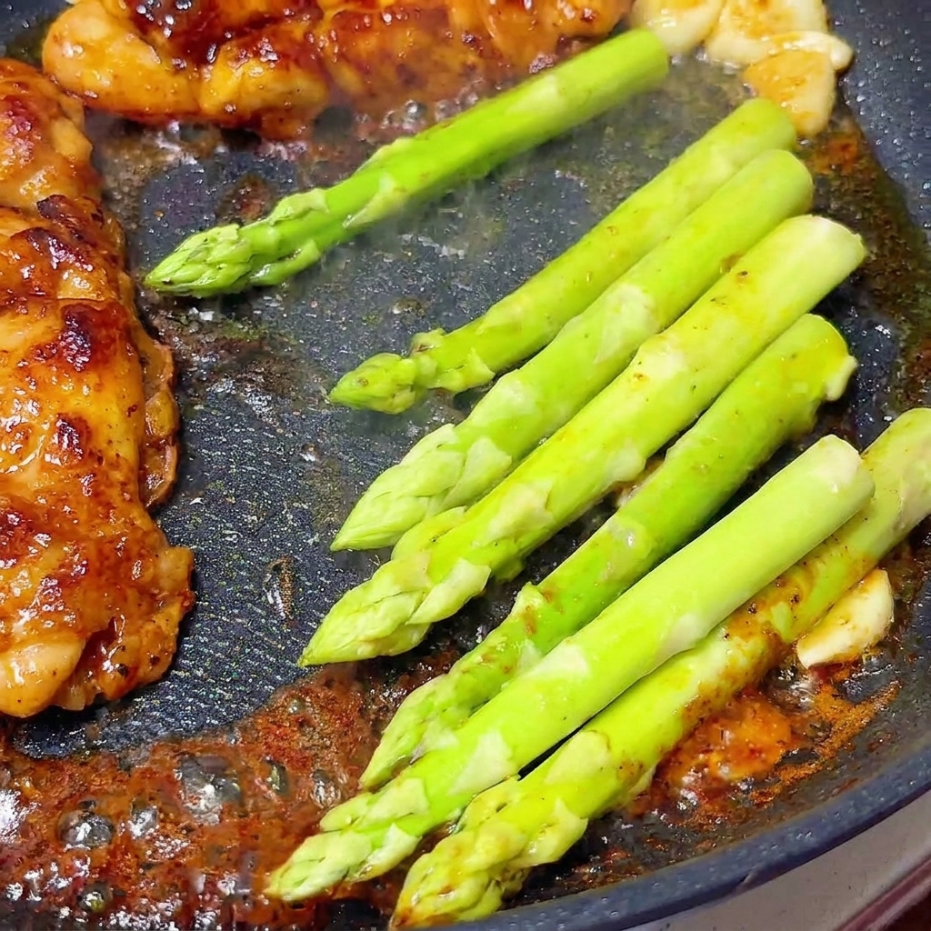 Asparagi freschi e di un verde brillante che cuociono a fuoco lento accanto ai petti di pollo glassati in una padella.
