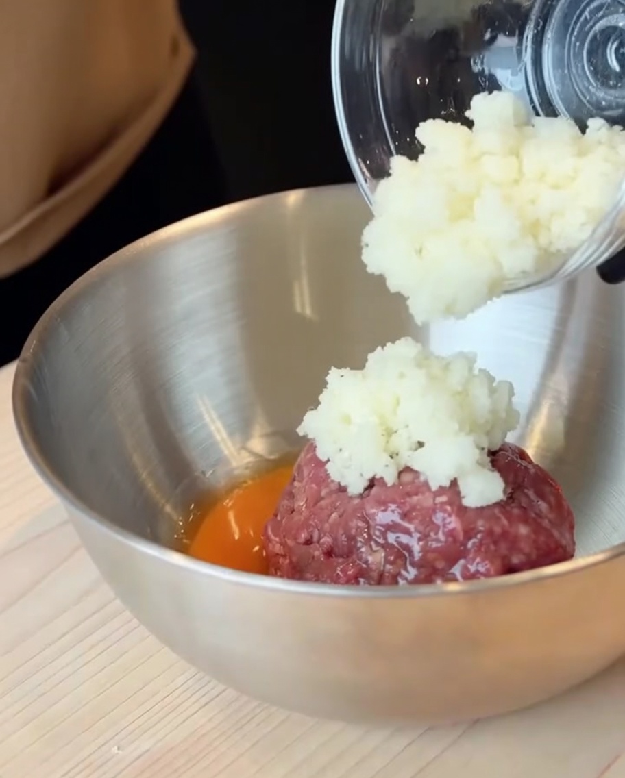 Carne macinata cruda e un uovo rotto in una grande ciotola di acciaio inossidabile con laggiunta di cipolle a dadini.