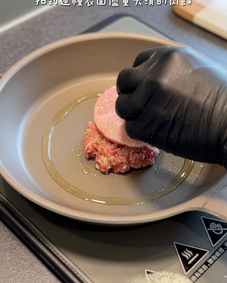 Una polpetta di hamburger cruda che viene delicatamente adagiata in una padella calda e oleata usando una mano con guanto nero e una spatola.