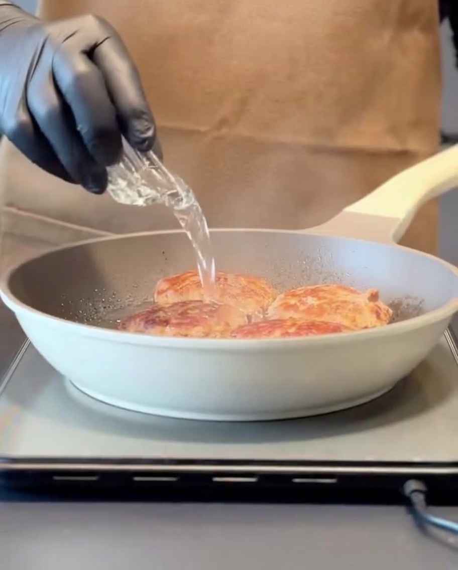 Acqua versata in una padella calda contenente polpette di carne rosolate, creando vapore prima di coprire con un coperchio.