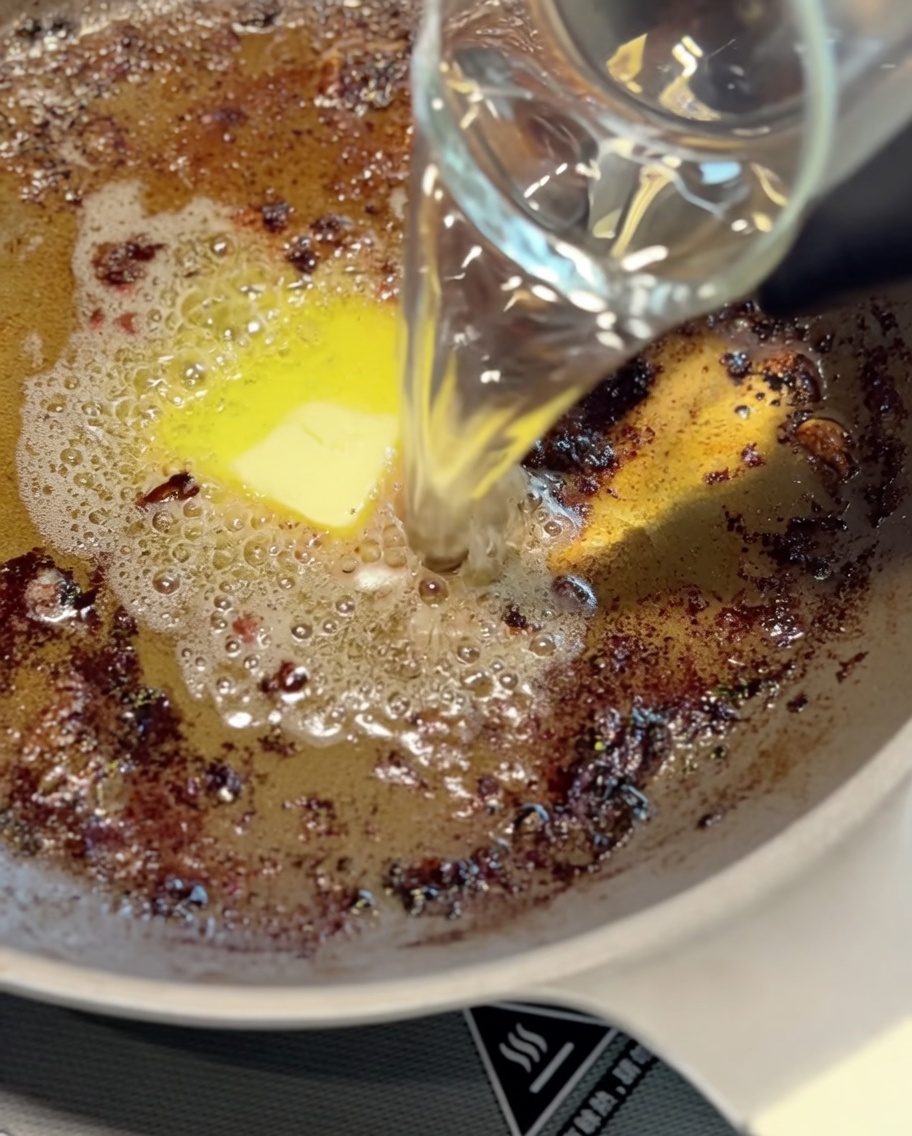 Un mestolo di metallo che mescola i succhi di cottura rosolati e acqua in una padella per formare la base della salsa.