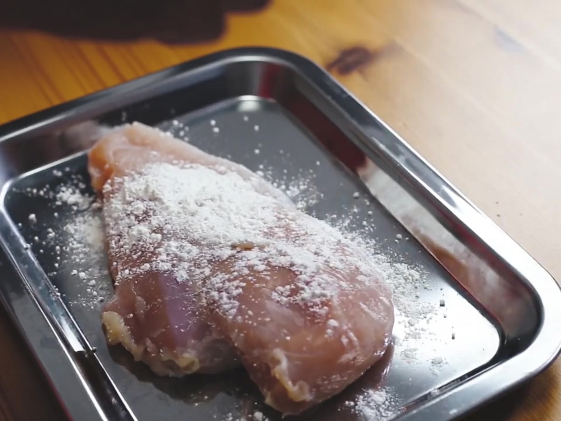Petto di pollo crudo ricoperto di farina su un vassoio di metallo.