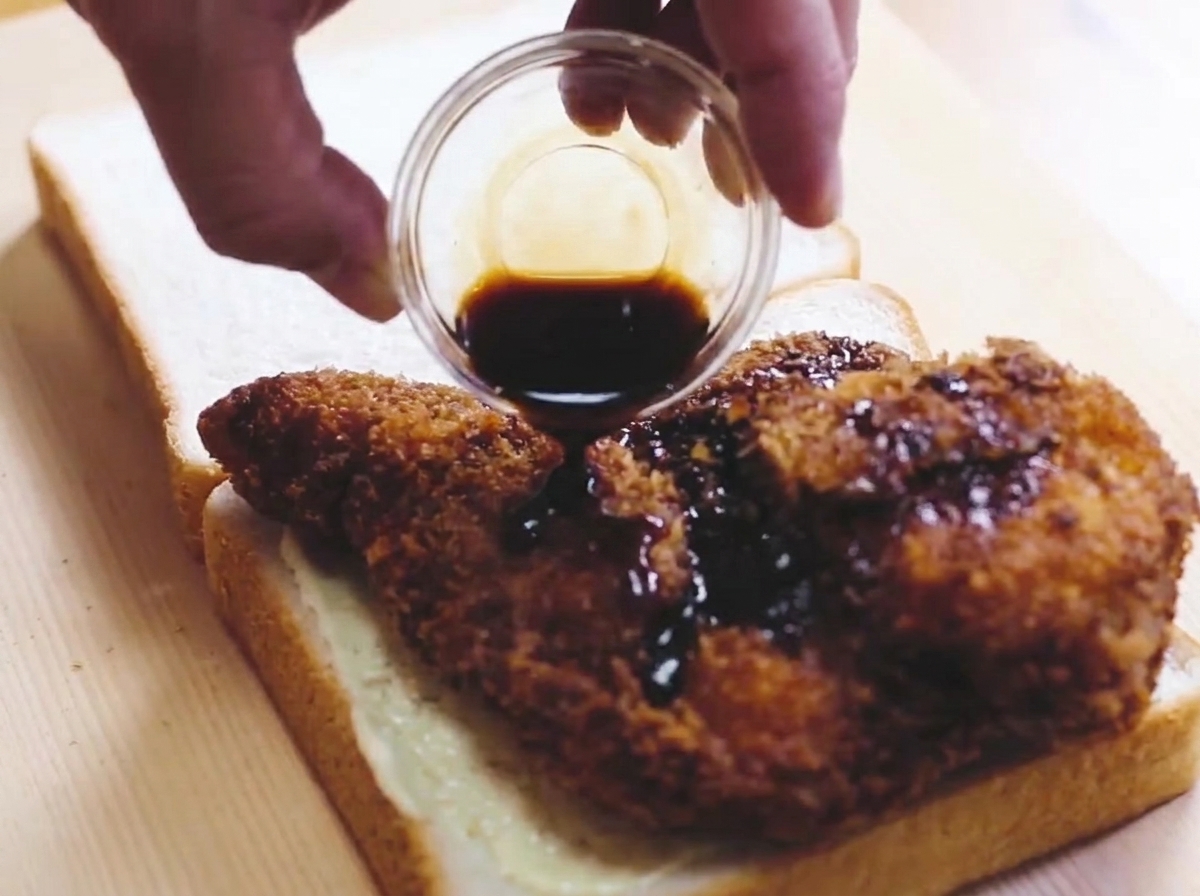 Una cotoletta di pollo fritto ben dorata appoggiata su una fetta di pane bianco e guarnita con salsa scura.