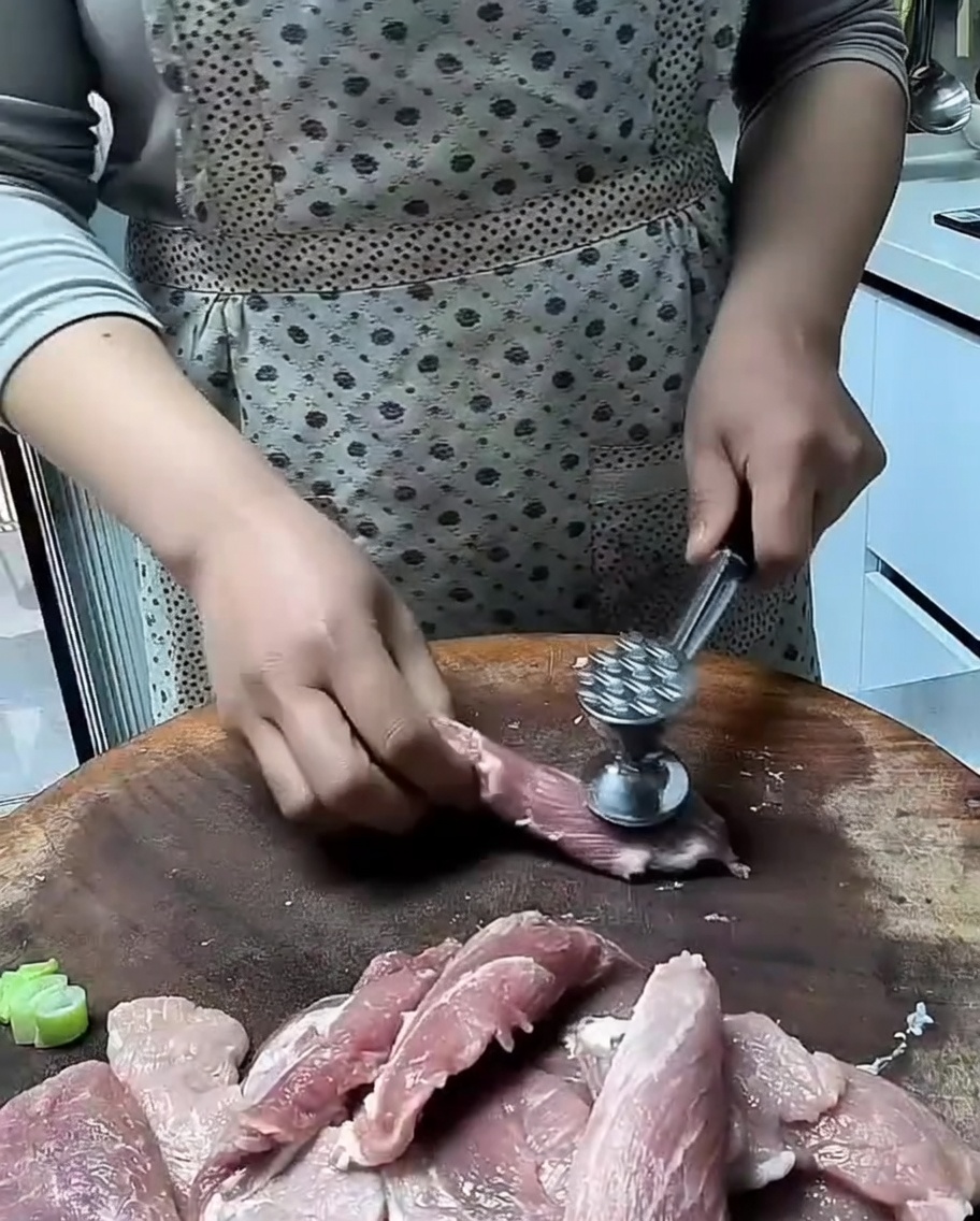 Un batticarne in metallo che intenerisce fette sottili di maiale crudo su una superficie in legno.