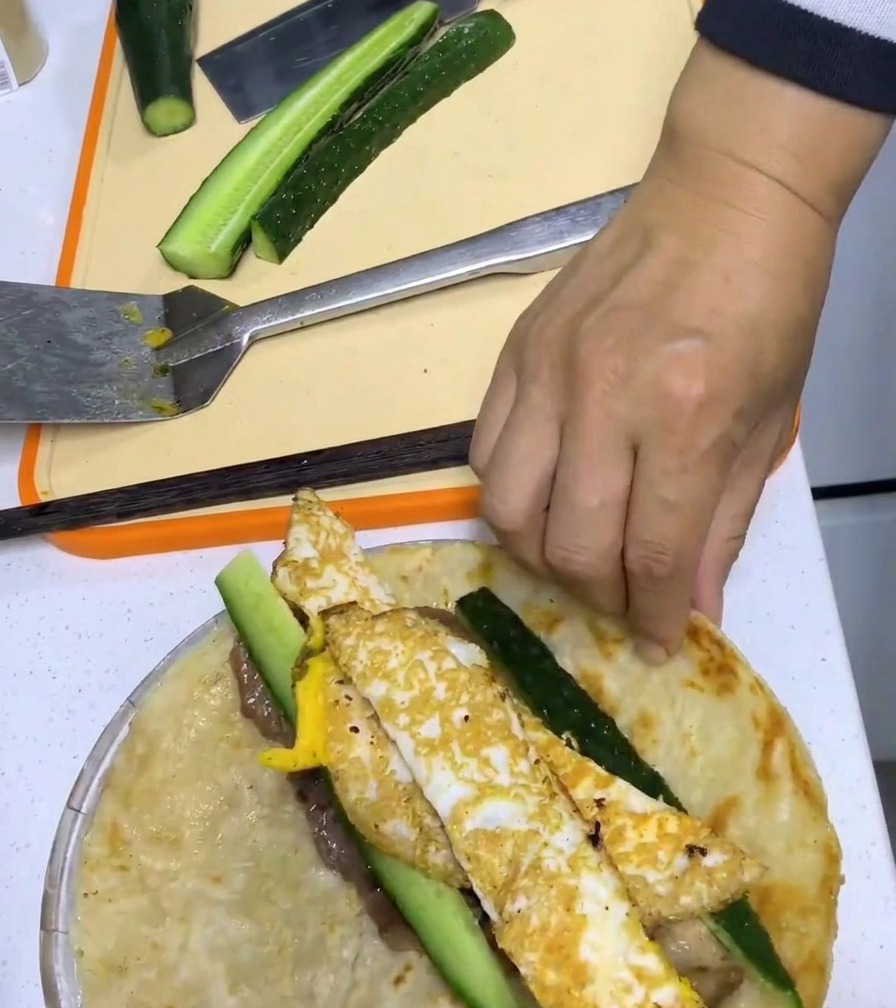 Una mano che arrotola strettamente un pancake allo scalogno attorno a maiale, uovo e cetriolo.