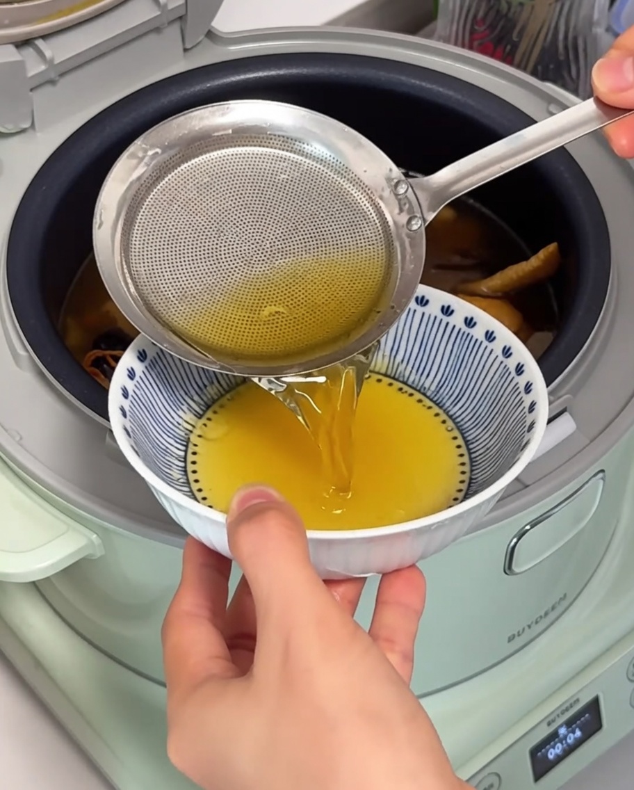 Primo piano di un colino a maglie fini che rimuove il grasso di pollo limpido e dorato dalla superficie della zuppa.