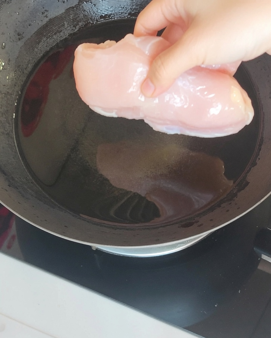 Un petto di pollo immerso in acqua bollente in un wok insieme ad aromi come zenzero e cipollotto.