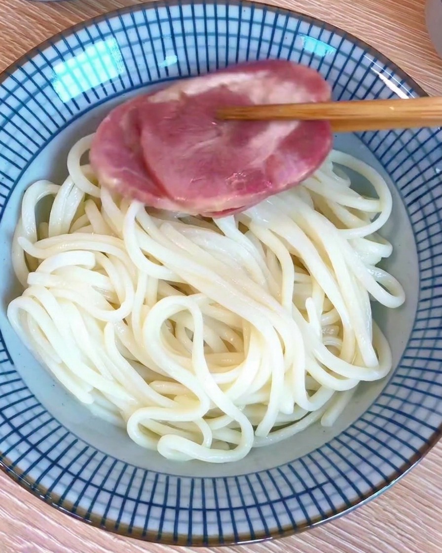 Una ciotola piena di noodles cotti guarniti con una fetta di maiale chashu, con bacchette che tengono la fetta di carne.