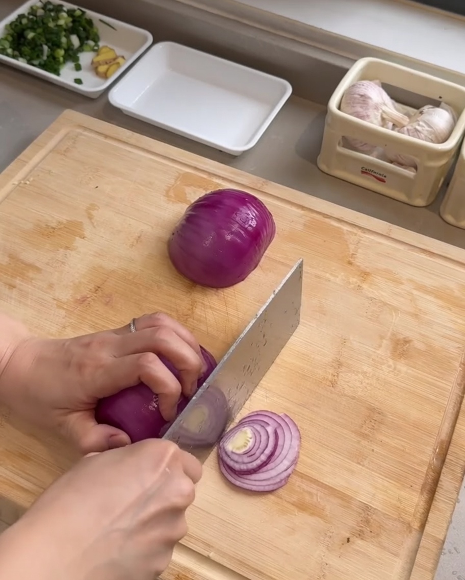 Un coltello da chef che affetta una cipolla rossa in strisce sottili su un tagliere di legno.