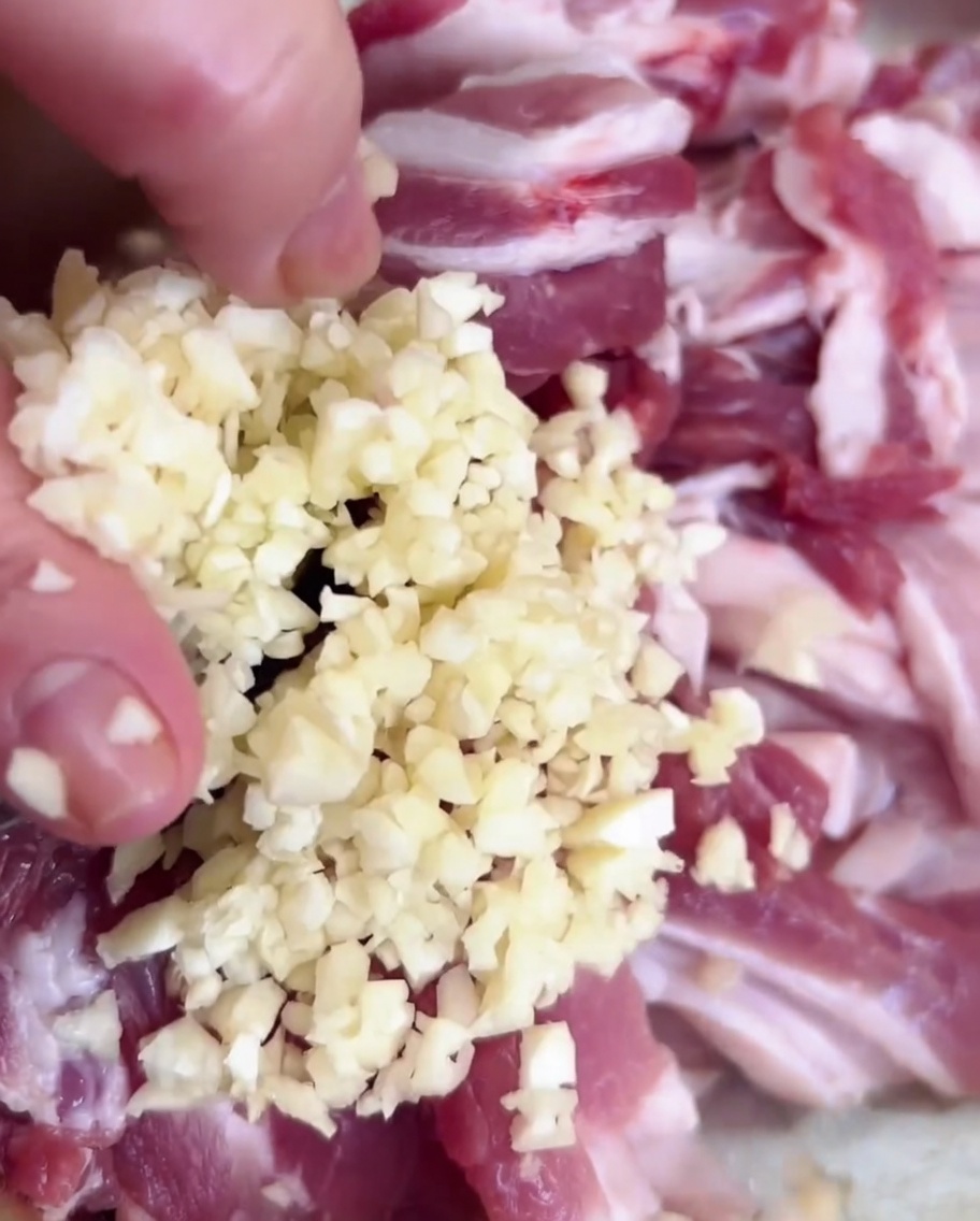 Aglio tritato versato da una mano in una ciotola di metallo contenente maiale a fette.