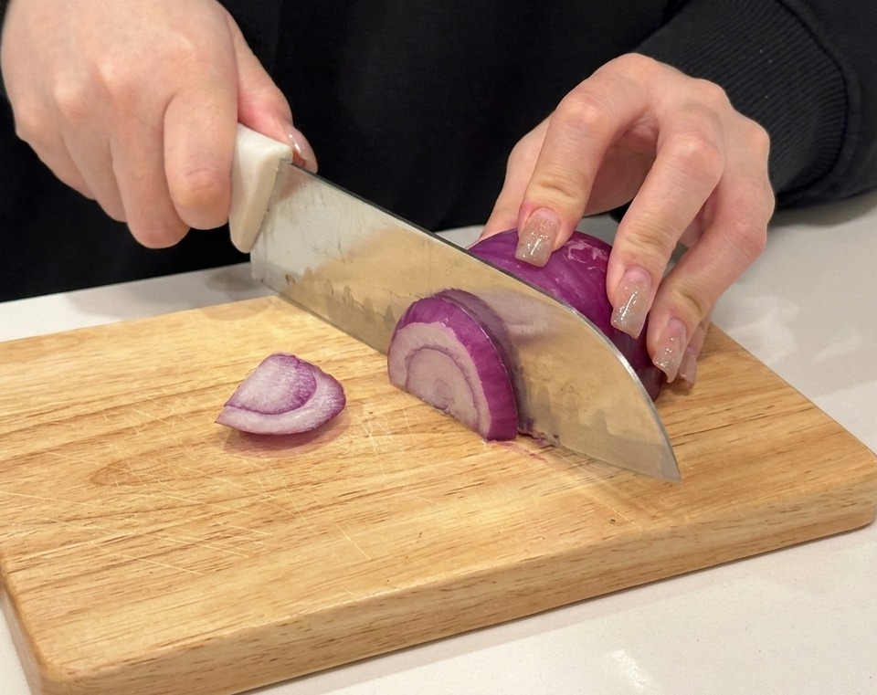 Mani che affettano una cipolla rossa su un tagliere di legno.