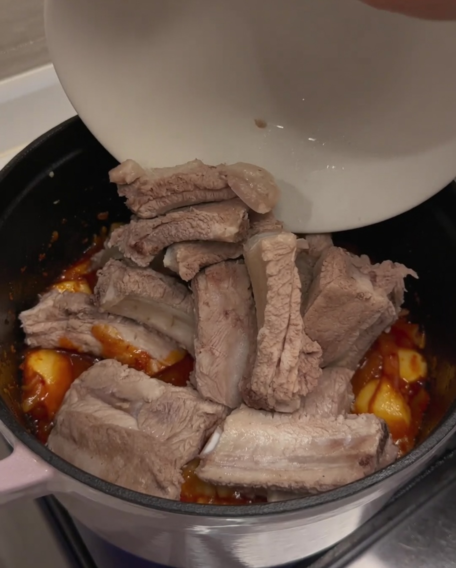 Costine di maiale precedentemente sbollentate aggiunte da una ciotola bianca nella pentola contenente la base di kimchi condita.