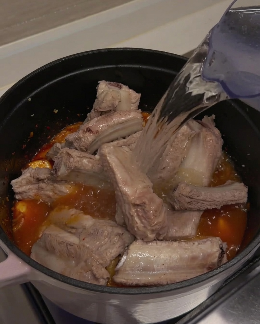 Acqua pulita versata da una brocca in una pentola, coprendo completamente le costine di maiale e la miscela di kimchi.