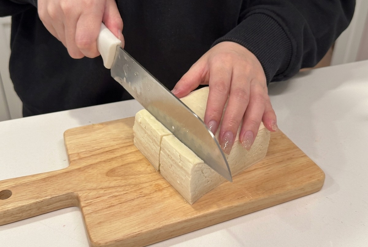 Dettaglio di un coltello che taglia un blocco di tofu compatto a cubetti su un tagliere di legno.
