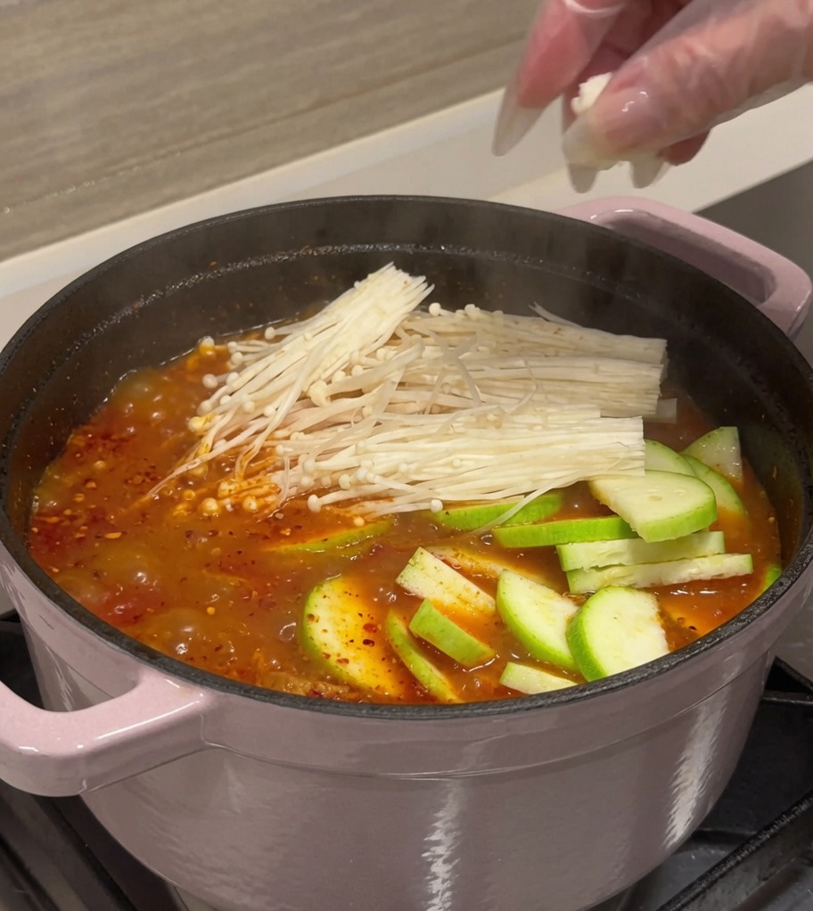 Zucchine a fette, cubetti di tofu e funghi enoki aggiunti a una pentola di brodo di kimchi rosso bollente.