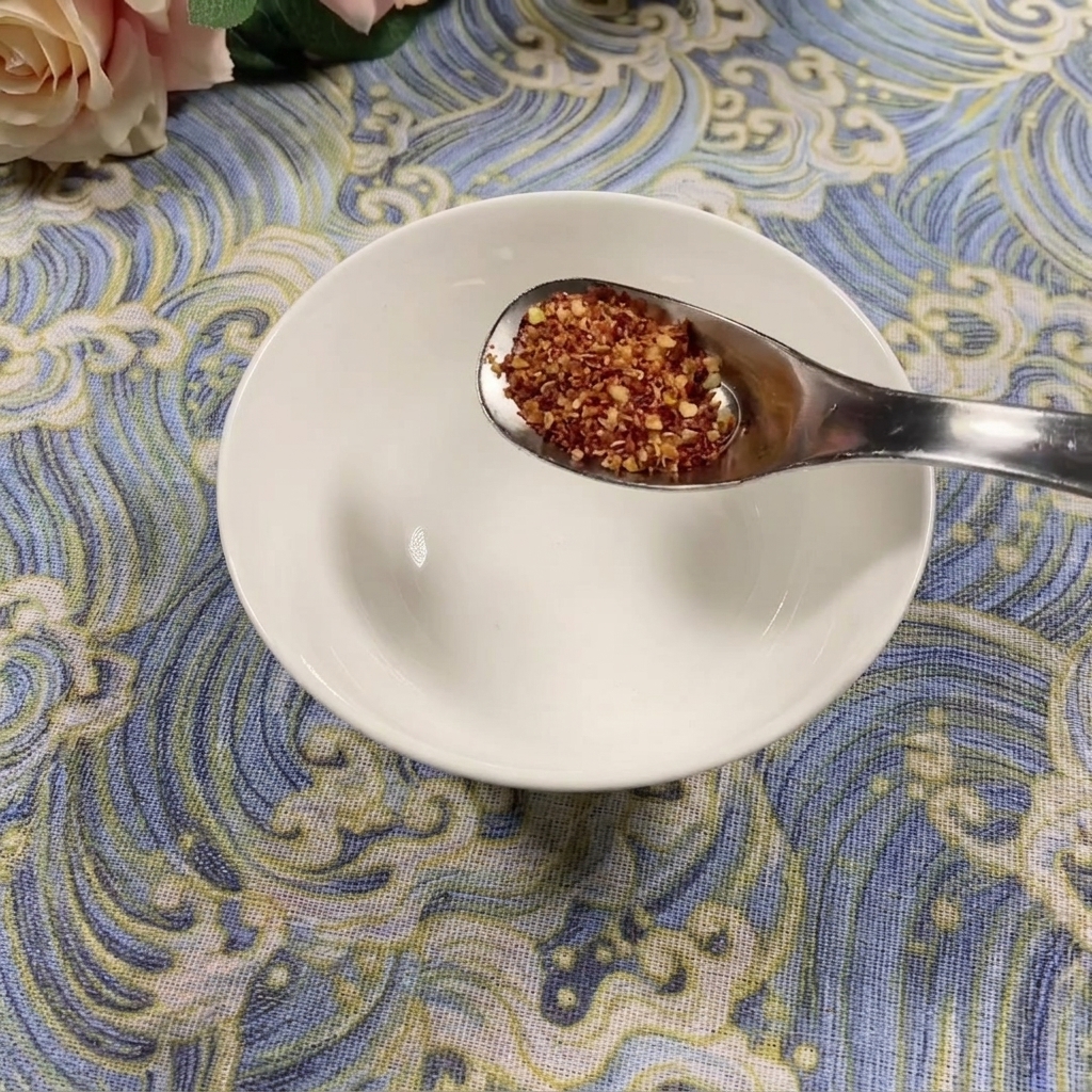 Un cucchiaio di peperoncino in polvere a scaglie grosse tenuto sopra una ciotola bianca vuota su una superficie blu a motivi.