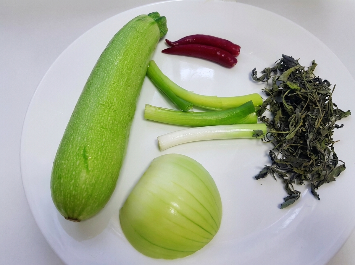 Ingredienti crudi tra cui una zucchina verde intera, cipollotti, cipolla bianca, foglie di perilla selvatica e peperoncini rossi sono disposti su un piatto bianco.