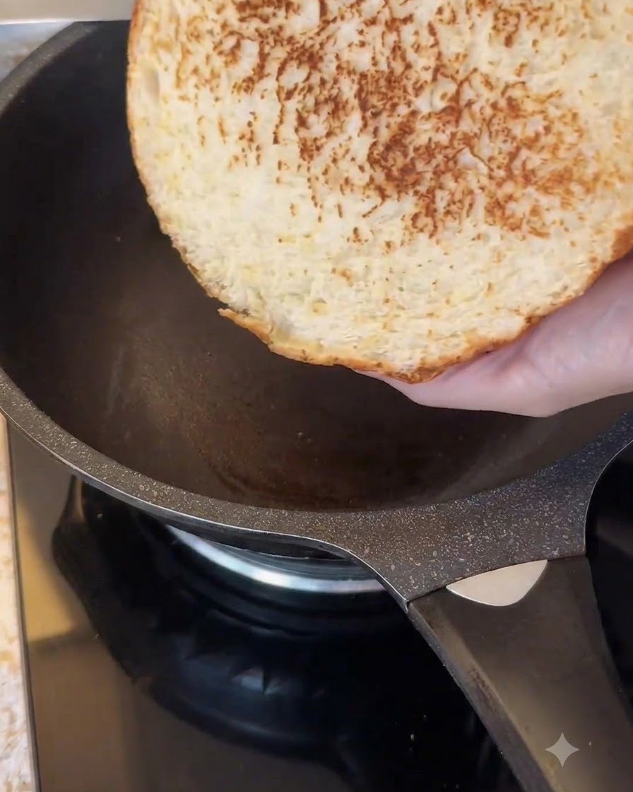Il lato tagliato di un grande panino per hamburger che viene tostato in una padella antiaderente scura.