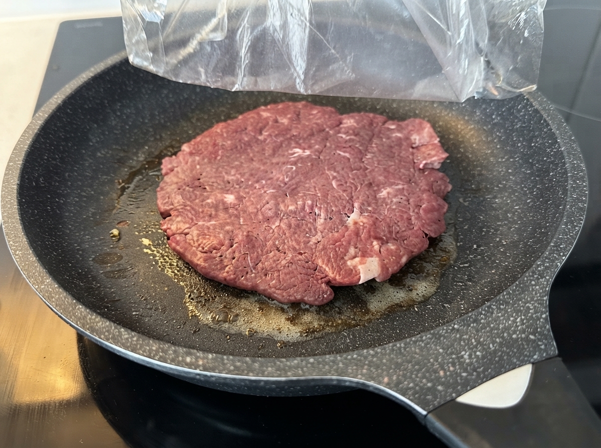 Un grande patty di manzo crudo che cuoce in una padella antiaderente unta sopra un piano cottura.