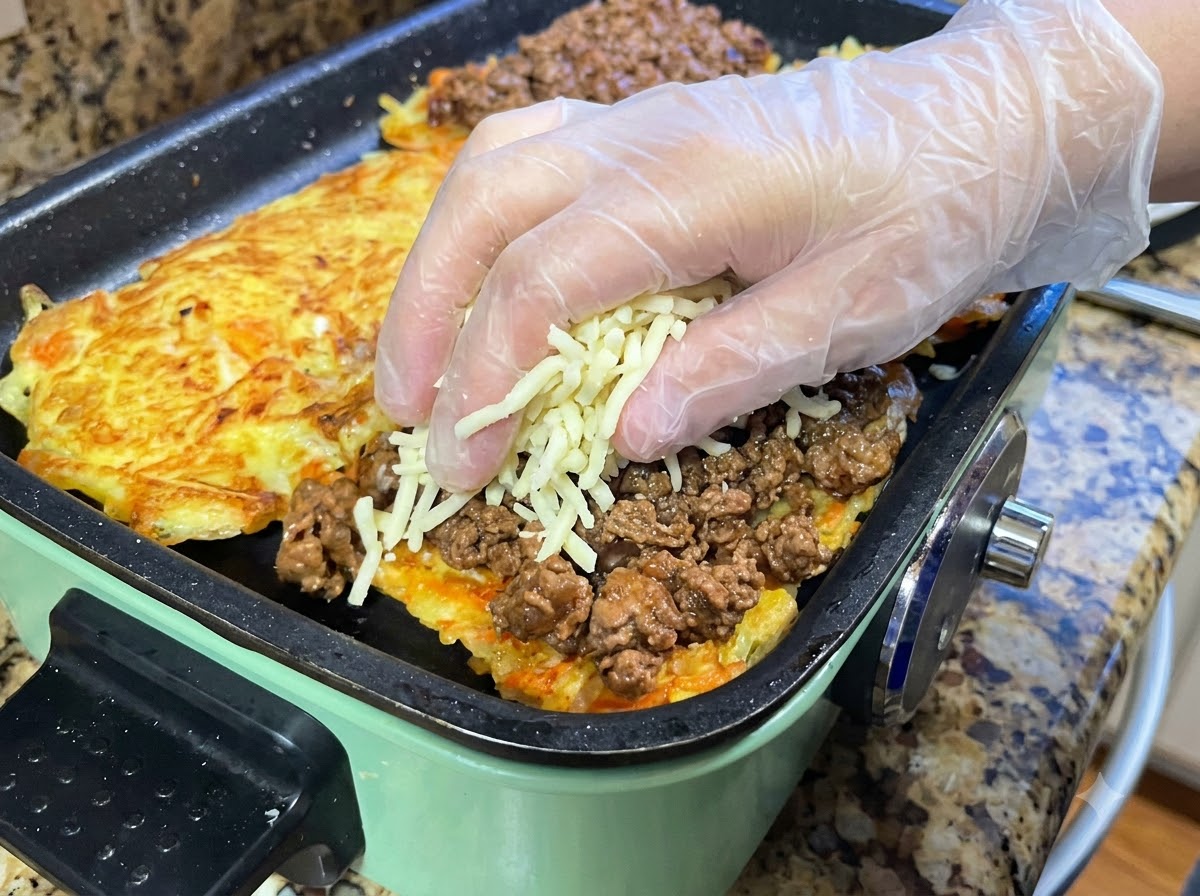 Mani con guanti protettivi che distribuiscono una manciata di mozzarella grattugiata sopra uno strato di salsa di carne cotta stesa su una frittata di patate.