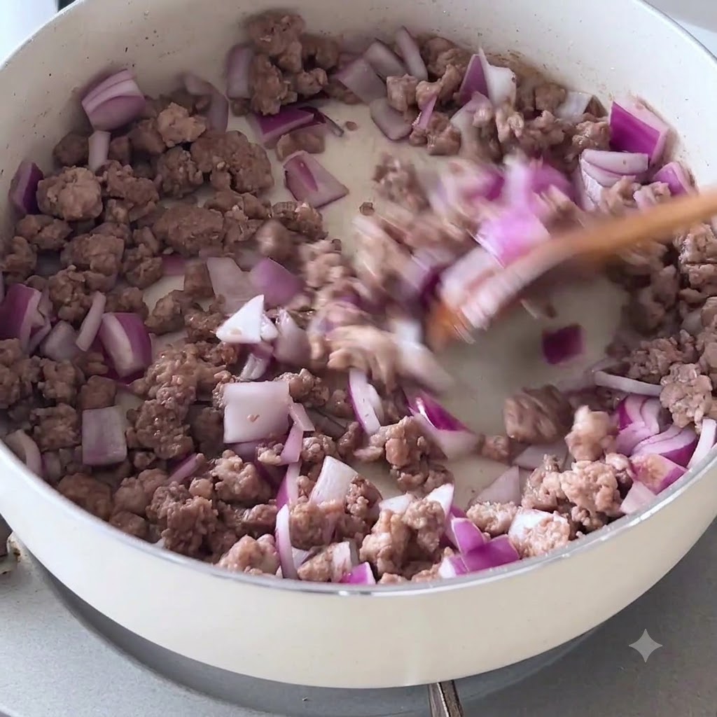 Cipolle rosse a cubetti e carne rosolata mescolati con una spatola di legno in padella.