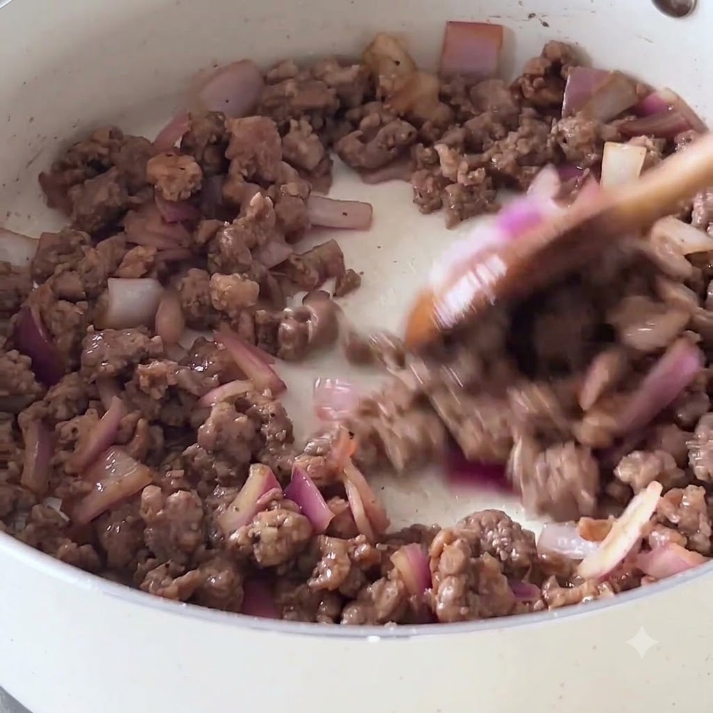 Carne macinata e cipolle rosse saltate in una padella antiaderente bianca con un cucchiaio di legno.