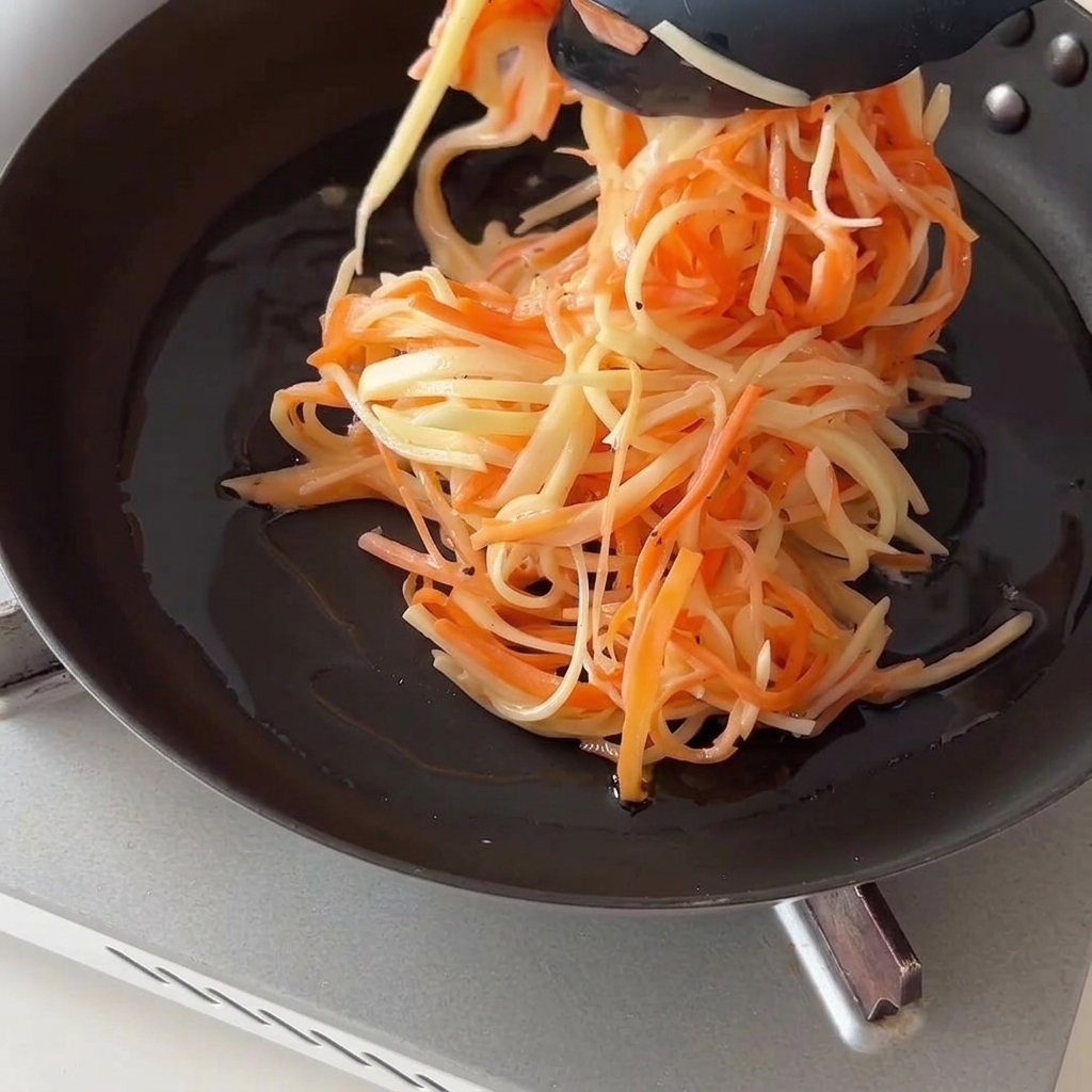 Uso di pinze da cucina nere per mettere il composto di patate e carote in una padella oliata.