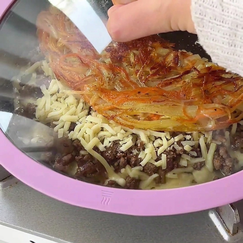Un coperchio di vetro posizionato su una padella contenente crosta di patate, carne macinata e formaggio che fonde.
