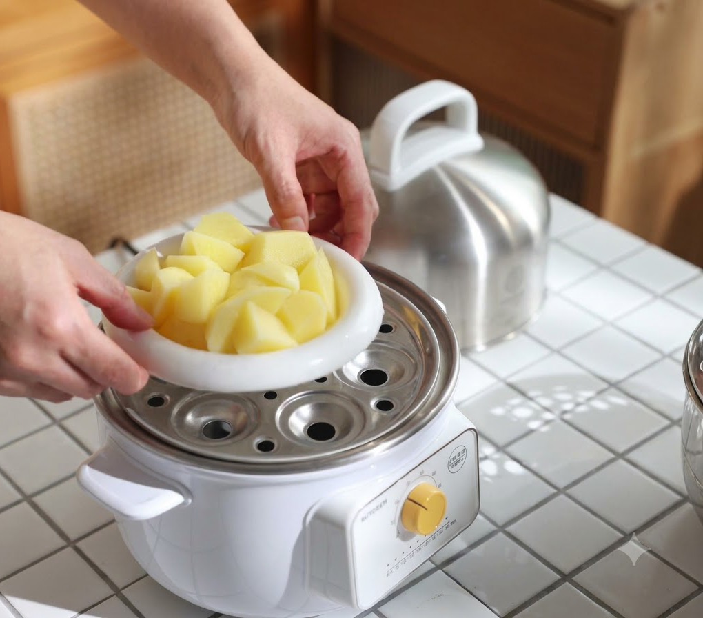 Un piatto bianco pieno di pezzi di patate inserito in una pentola per la cottura al vapore fino a renderle tenere.
