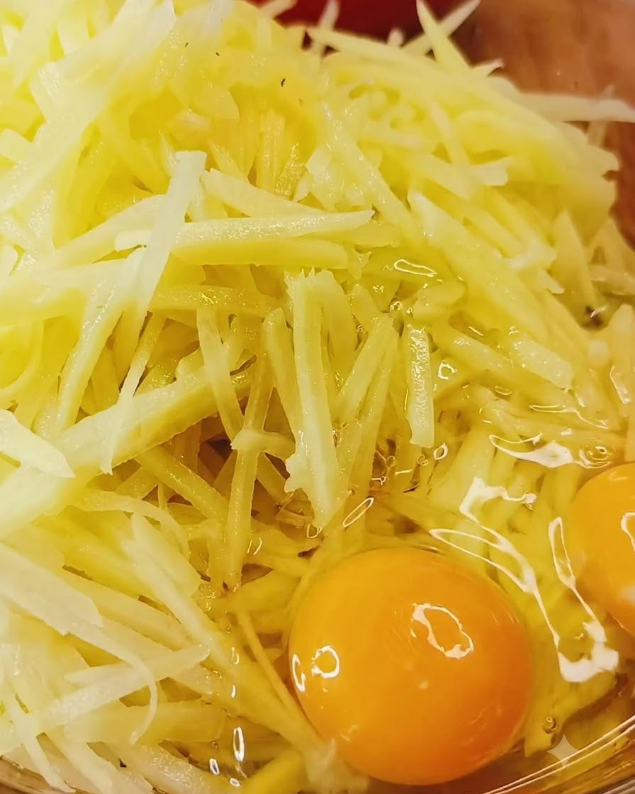 Uova intere rotte direttamente sopra una ciotola di patate crude grattugiate e condimenti.
