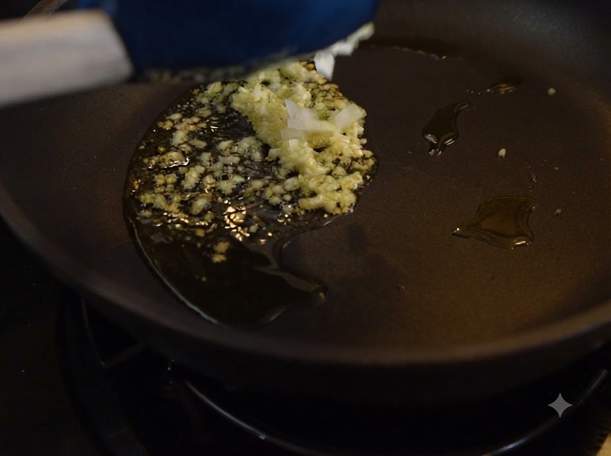 Aglio tritato e cipolle in cottura nellolio allinterno di una padella scura.