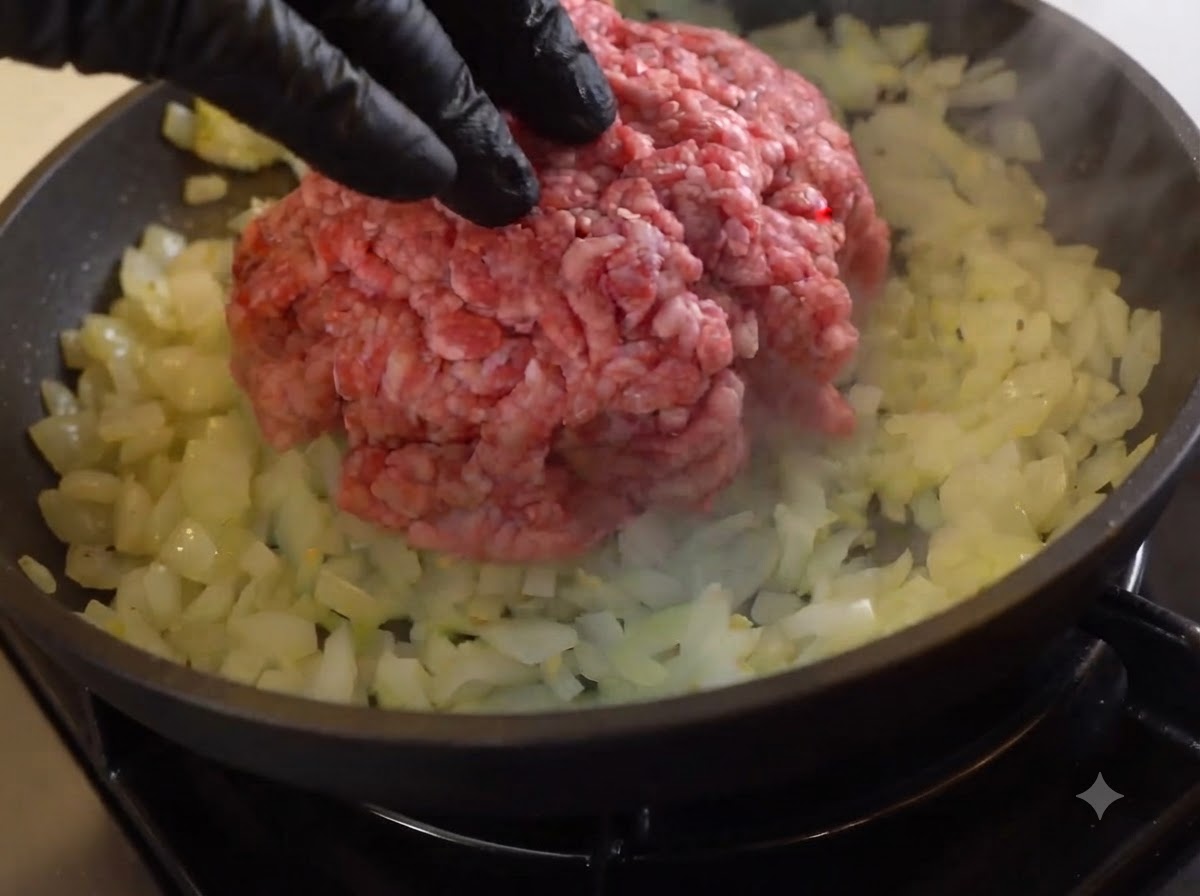 Un mucchio di carne macinata cruda adagiata sopra le cipolle soffritte in una padella.