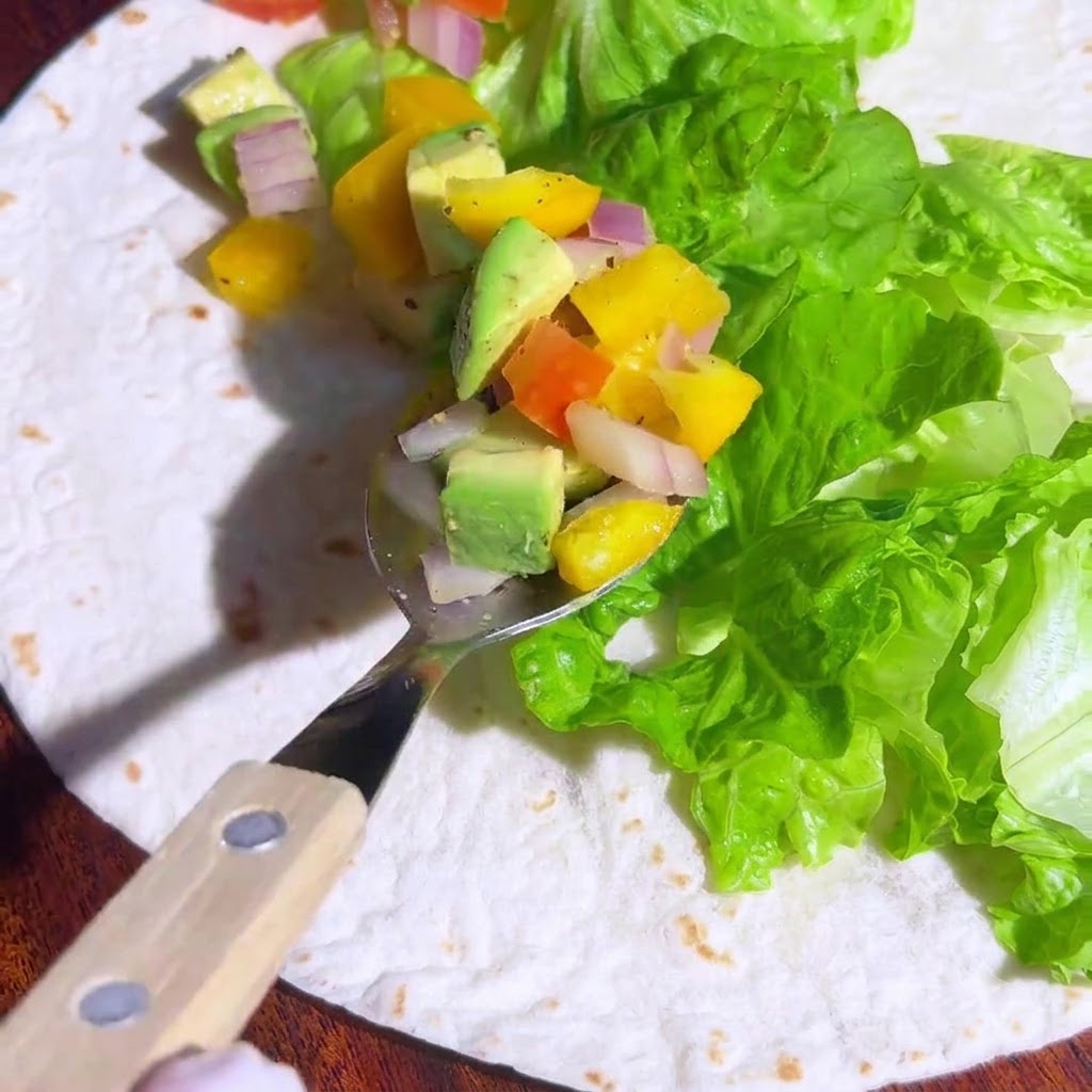 Un cucchiaio pieno di salsa colorata a base di avocado e verdure, posizionato sopra la lattuga su una tortilla.