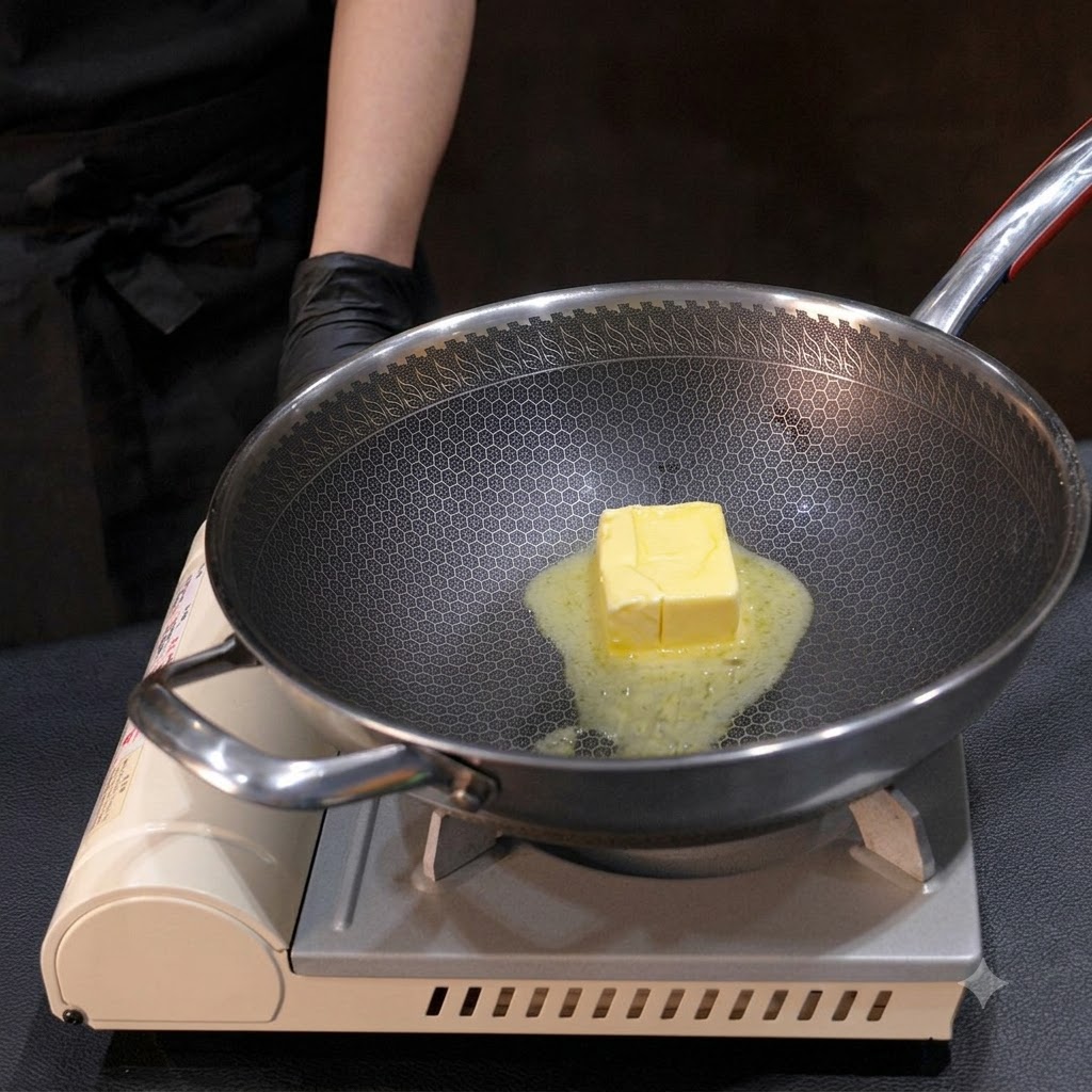 Un panetto di burro giallo che si scioglie in un wok di metallo, mescolato con una piccola frusta sul fuoco.