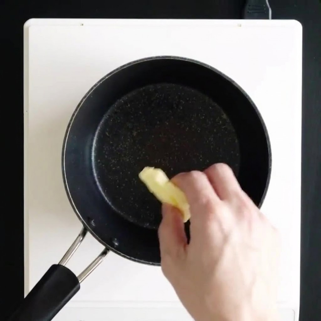 Una mano che aggiunge pezzi di burro in una casseruola nera su un piano cottura bianco per farlo sciogliere.