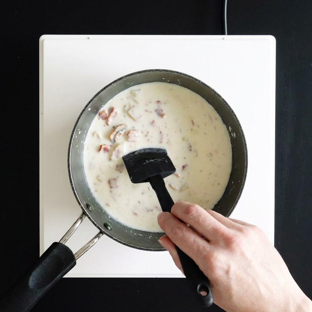 Una mano che mescola una zuppa bianca densa e cremosa in una casseruola usando una spatola in silicone nero.